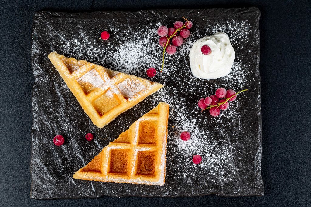Triangular waffles with currant, powdered sugar and whipped cream on a black background, top view