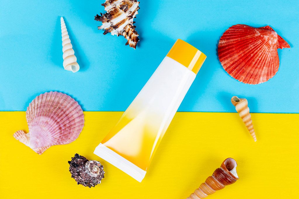 Tube of sunscreen with shells on a yellow and blue background. Beach vacation concept, top view