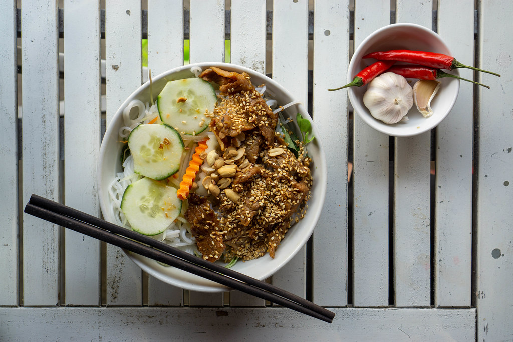 Vietnamesisches Gericht Bun Thit Nuong mit Reisnudeln, gegrilltem Schweinefleisch, Sesam, Gurken, eingelegten Karotten und Ernüsssen von oben auf einem weißen Holztisch fotografiert