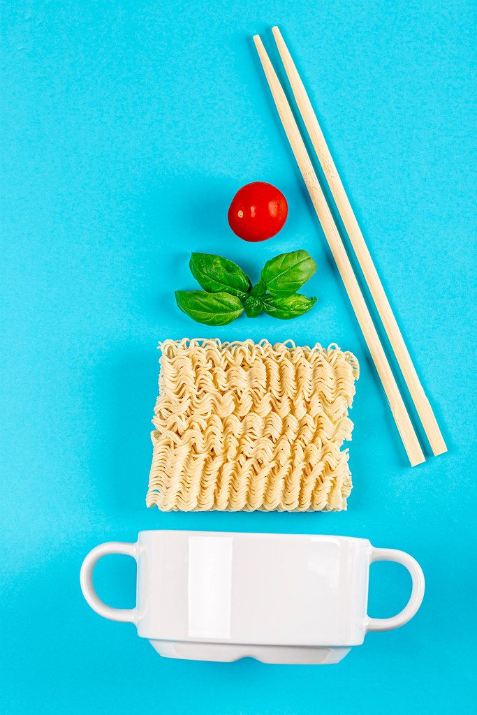White tureen on a blue background with ingredients for a quick lunch. Raw instant noodles