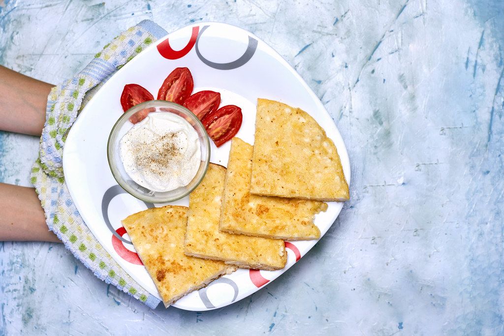 Woman holding rice-flour based khachapuri with a bowl of sour cream and fresh tomato slices
