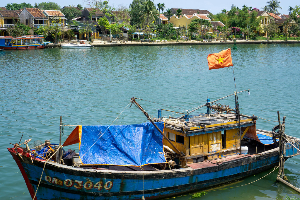 Wooden Fishing Boat with Rain Cover and Vietnamese Flag on a River with Houses and Palm Trees in the Background in Hoi An, Vietnam