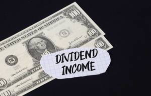 "Dividend income" Text auf kariertem Zettel mit zwei 100-Dollar-Scheinen vor schwarzem Hintergrund