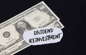 "Dividend reinvestment" Text auf kariertem Zettel mit zwei 100-Dollar-Scheinen vor schwarzem Hintergrund