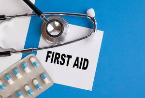 "First Aid" / Erste Hilfe, geschrieben auf blauem Ärzteordner, neben Medikamenten und Stethoskop