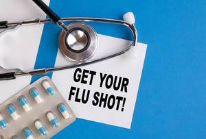 "Get your Flu Shot!" / Geh zur Grippeschutzimpfung, geschrieben auf blauem Ärzteordner, neben Medikamenten und Stethoskop