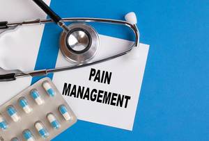"Pain Management" / Schmerzbehandlung geschrieben auf blauem Ärzteordner, neben Medikamenten und Stethoskop