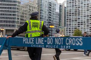 A Chicago police officer stands next to a large blue barrier with "police line - do not cross. Chicago Police Dept" written on it