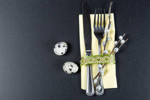 A fork and knife with a yellow napkin , willow branches and quail eggs on a black background. The concept of preparation for Easter