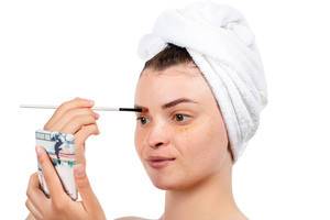 A girl with a white towel on her head dyes her eyebrows with a mirror in her hand