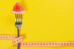 A piece of ripe grapefruit on a fork wrapped with measuring tape on a yellow background as a symbol of weight loss (Flip 2019)