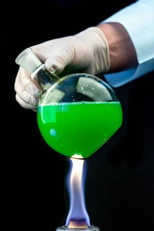 A round flask with a green solution in the hand of a scientist over a fire on a dark background (Flip 2020)