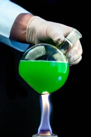 A round flask with a green solution in the hand of a scientist over a fire on a dark background