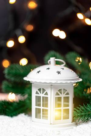 A white christmas lantern with lit candle and tree branches in front of dark background with lights