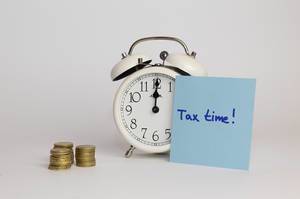 Alarm Clock standing next to a stack of coins and a note saying tax time on white background