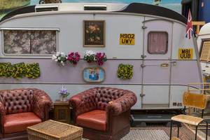 Alter Wohnwagen mit Blumen & Bildern von Lady Di und Prinz Charles als Hostelzimmer mit Wohnzimmerflair & Vintagesessel aus rotem Leder