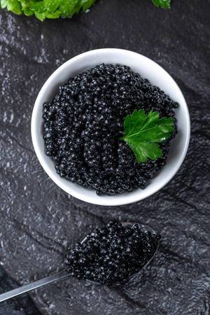 Ansicht von obenauf schwarzen Kaviar auf einem Löffel und in einer weißen Schale auf schwarzer Schieferplatte