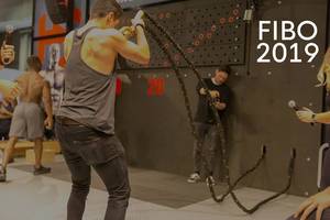 Athlet und Sportmessebesucher trainiert am Battlerope vor Zuschauern, neben dem Bildtitel "Fibo 2019"