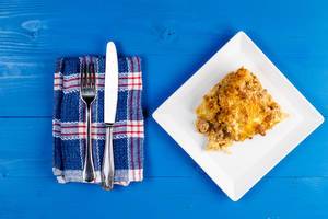 Auflauf mit Kartoffeln und Gehacktes auf weißem Teller neben Küchentuch und Besteck auf blauem Holztisch