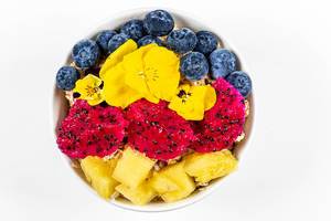 Aufnahme von oben: frische Stücke Pitahaya, Ananas, Blaubeeren und Blumen mit Haferflocken in einer Schüssel vor weißem Hintergrund