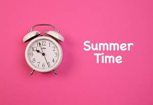 Aufschrift "Summer Time" - Sommerzeit, erinnert an die Zeitumstellung, neben einem weißen Vintage-Wecker, auf pink-blauem Hintergrund