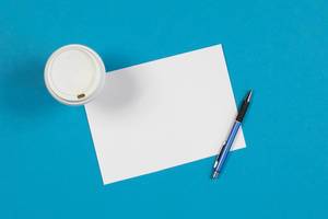 Aufsicht - Ein Becher Kaffee mit einem leeren Blatt Papier und Stift auf blauem Hintergrund