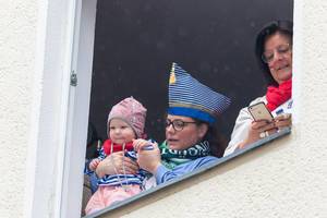 Baby und Mama in passendem Outfit in blau-weiß verfolgen den Rosenmontagsumzug am Fenster. Neben ihnen eine andere Frau mit dem Handy