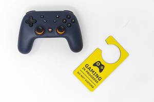 Basic equipment for gaming: Stadia video game controller and yellow door hanger "Gaming in progress - do not disturb", seen from above with white background