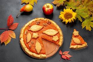 Beautiful decorated pumpkin pie on black background with autumn leaves, apple and sunflower flower