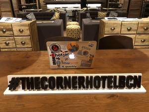 Beklebtes Macbook auf einem Holztisch in der Lounge vom The Corner Hotel, hinter dem Hashtagbanner #TheCornerHotelBCN