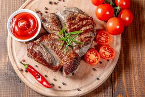 Blick von oben auf zarte Steaks mit Soße, Tomaten, Pfeffer, Chilischote und Rosmarin auf Küchenbrett