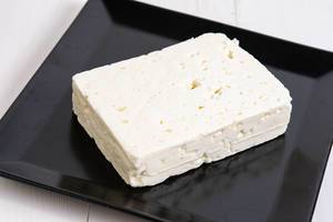 Block eingelegter weißer Käse auf schwarzem, quadratischem Teller auf weißem Holztisch