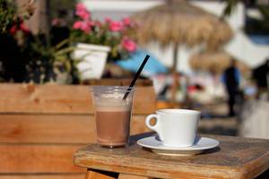 Bokeh Photo of Ceramic Cup of Coffee and Plastic Cup with Cocoa Hot Chocolate on Wooden Table with Plants in the Background