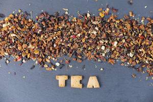 Buchstabenkekse formen das Wort "Tea" vor getrocknetem Tee, auf einem schwarzen Tisch