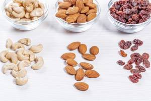 Cashews, almonds and raisins in glass bowls and scattered on a white wooden background