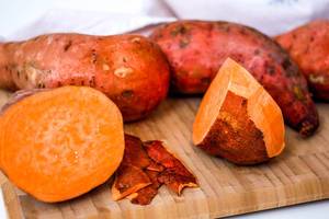 Close-up of a sweet potato cut in half and partially peeled, on a wooden chopping board with some more whole sweet potatoes