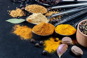 Close Up on spices on spoons - curcuma, curry, garlic, mustardseeds and barberries with nutmeg in front on black background