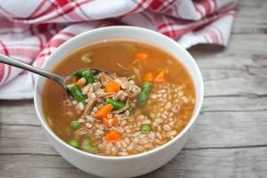 Close Up View of Bowl of Barley Soup with Beef, Pole Beans and Carrots with Spoon on Wooden Table