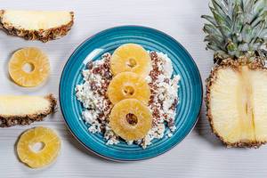 Close-up von Hüttenkäse mit Leinsamen und getrockneten Ananas Scheiben vor weißem Holzhintergrund mit frischen Ananas Stücken