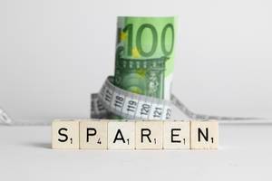 Das Wort Sparen auf Würfeln vor einem Euroschein mit Maßband auf weißem Hintergrund