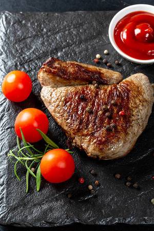 Delicious veal steak with tomatoes and spices on a black stone tray
