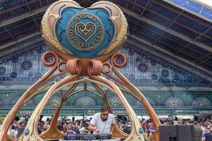 DJ spielt Musik innerhalb eines futuristischen DJ Stands während die Besucher in Schlangen warten um das Tomorrowland Festival zu betreten