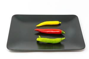 Drei kleine bunte Chilischoten in geld, rot und grün auf einem schwarzen Teller