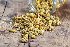 Dried chamomile flowers spill out of a glass jar on a wooden background