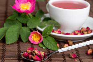 Dry tea rose buds in a spoon with a cup of tea and flowers behind