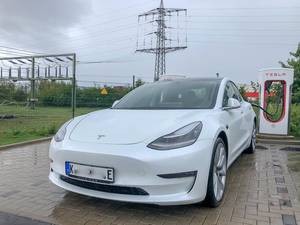 E-Mobilität: Weißer Tesla Model 3 lädt an einer Supercharger - Ladesäule bei Regenwetter
