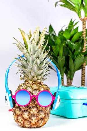 Eine Ananas trägt Sonnenbrille und Kopfhörer vor einem Hintergrund mit Koffer und Palmen