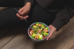 Eine auf dem Boden sitzende Person isst einen gesunden Salat mit Walnusskernen, Mikrogrün, Romanesco Kohl und Granatapfelkernen mit einer Gabel