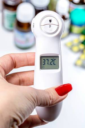 Eine Frau hält ein medizinisches Elektrothermometer in der Hand mit der Temperatur von 37,2°C und Arzneimittel im Hintergrund