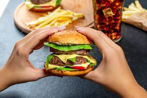 Eine Frau hält einen doppelten Burger in ihren Händen, mit Fast-Food im Hintergrund